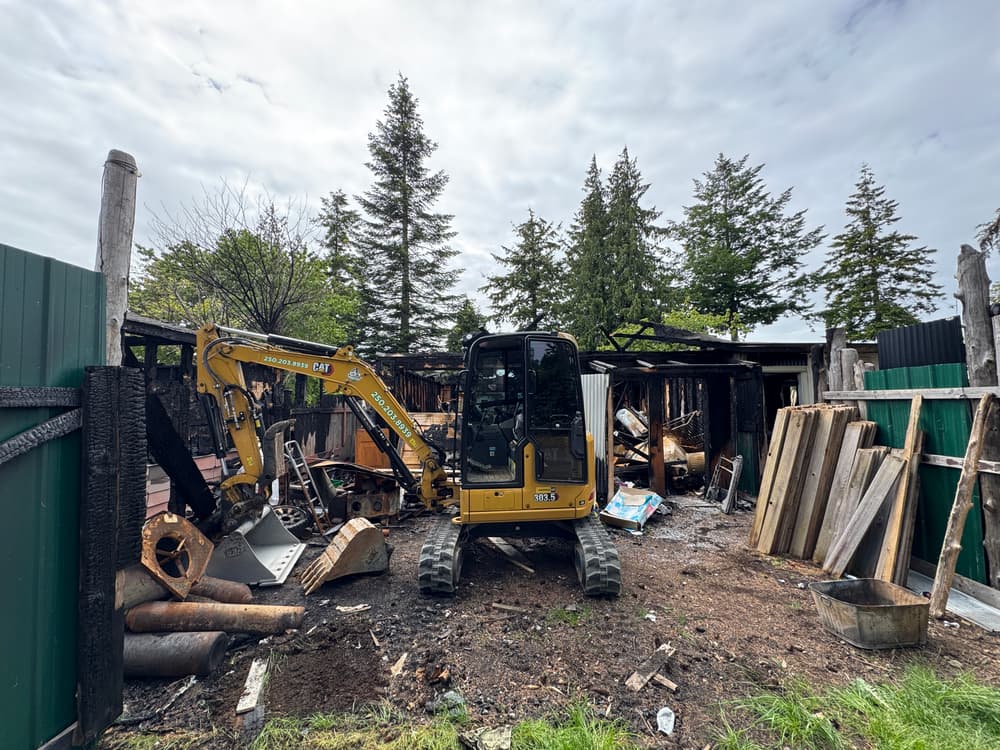 Excavator in fire-damaged area with charred structures and debris, surrounded by trees.