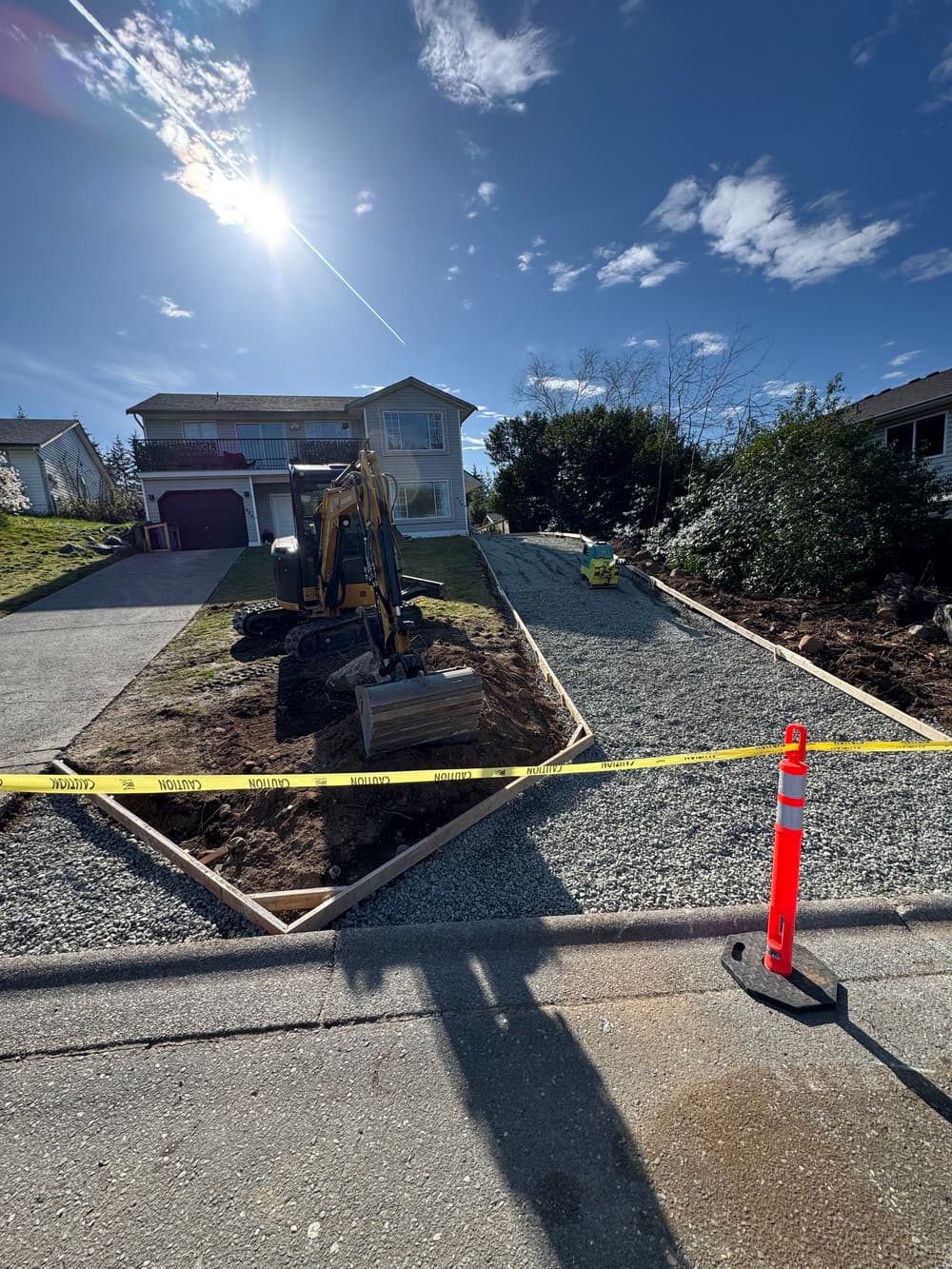 Excavator working on gravel driveway installation at residential property under sunny sky.