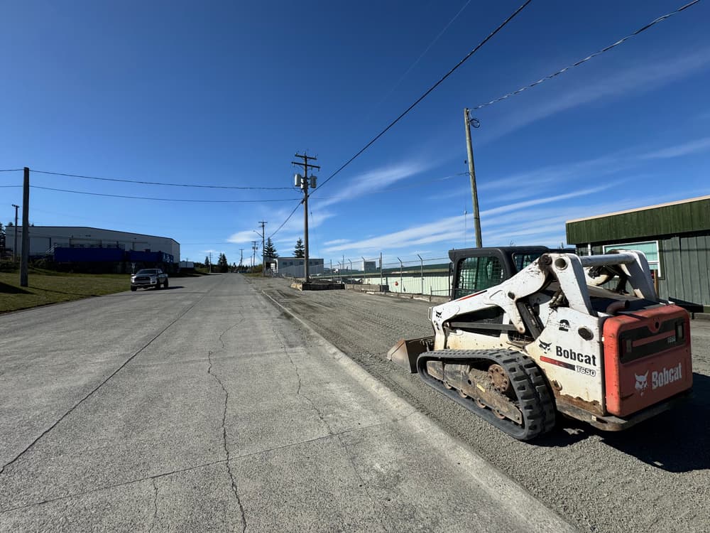 Bobcat skid steer parked on gravel road with utility poles and industrial buildings in background.