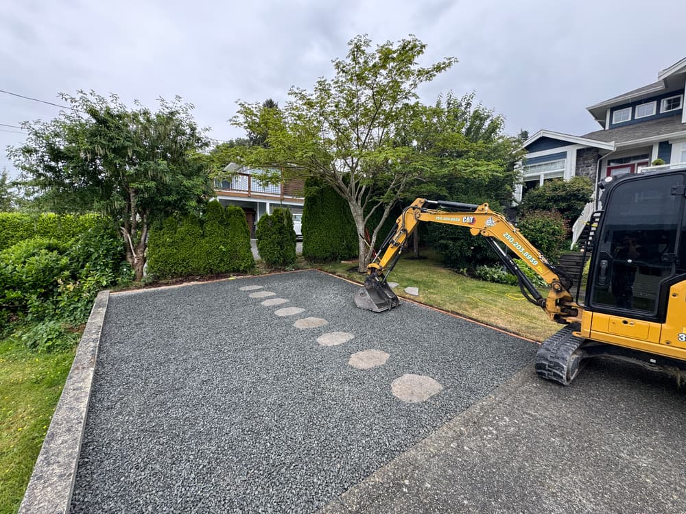 Excavator installing stepping stones in gravel driveway with greenery in background.