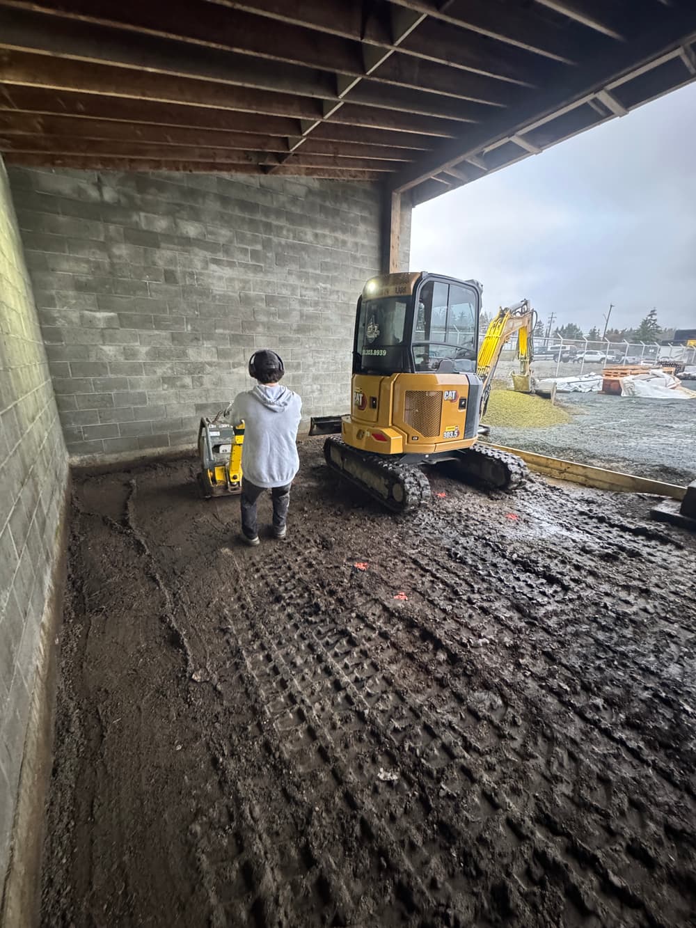 Construction worker operating a compact excavator on a muddy job site indoors.