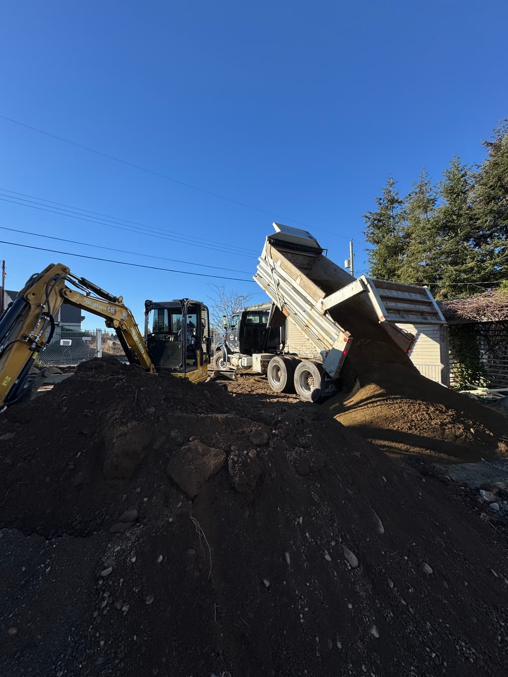 Excavators and dump trucks unloading soil at a construction site under a clear blue sky.
