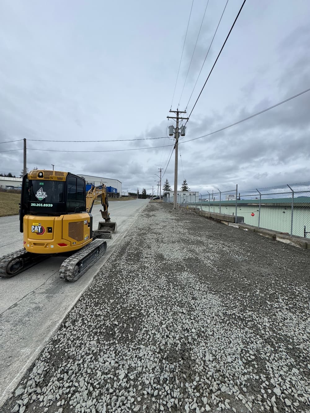 Yellow excavator working on a gravel road beside a fenced construction site under a cloudy sky.