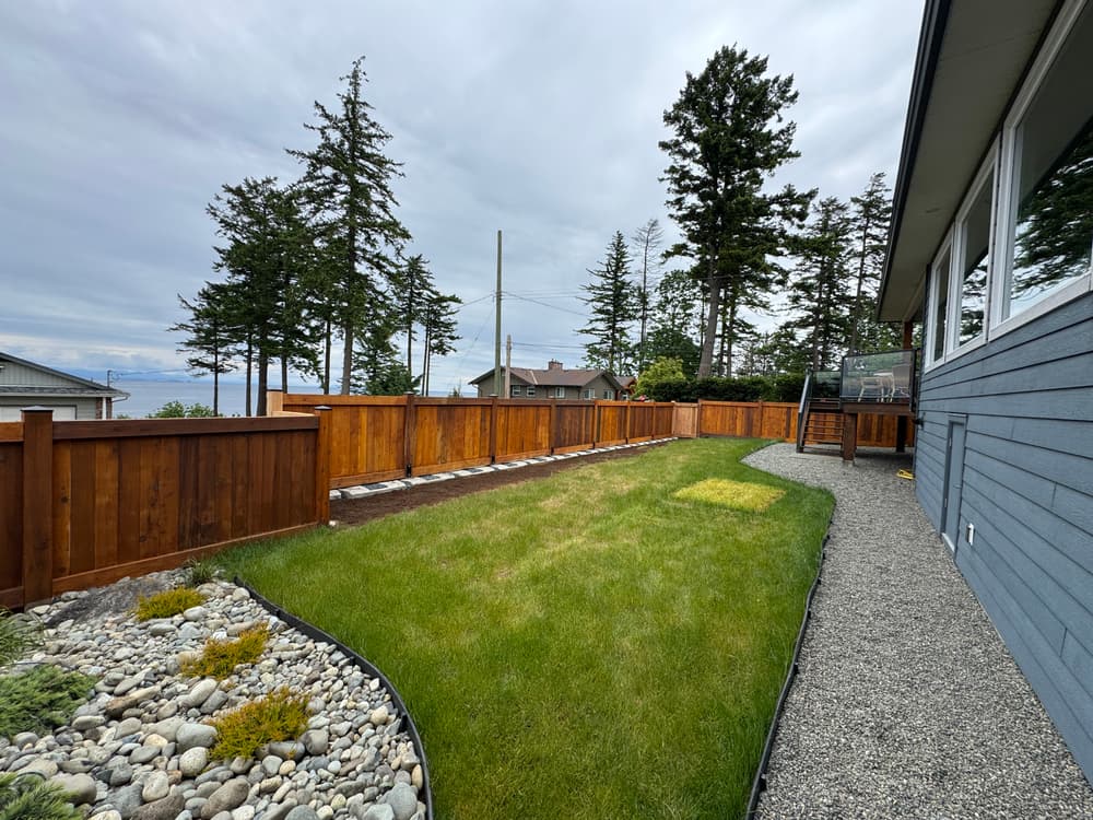 Lush green backyard with wooden fence and gravel path near coastal homes and trees.