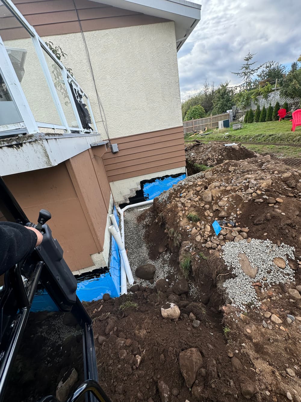 Excavation near a house with foundation work and drainage installation in progress.