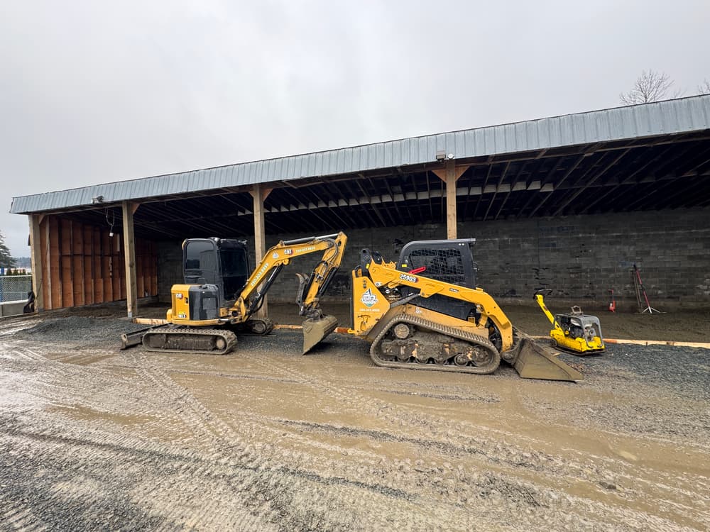 Two yellow construction excavators on a gravel site near a building under construction.