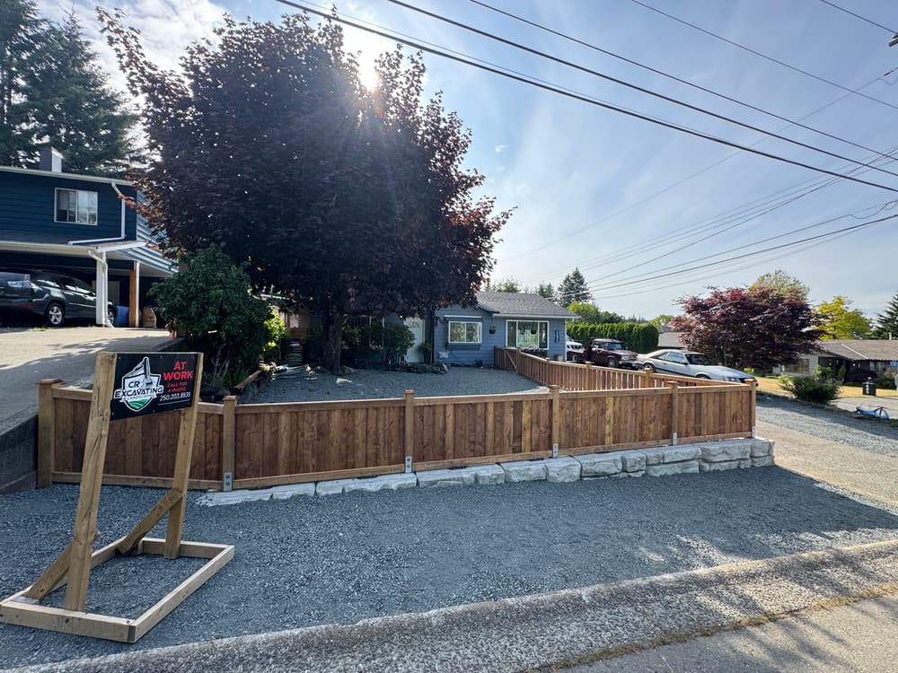 House with a fence, landscaped yard, and trees under sunny sky, with "At Work" sign visible.