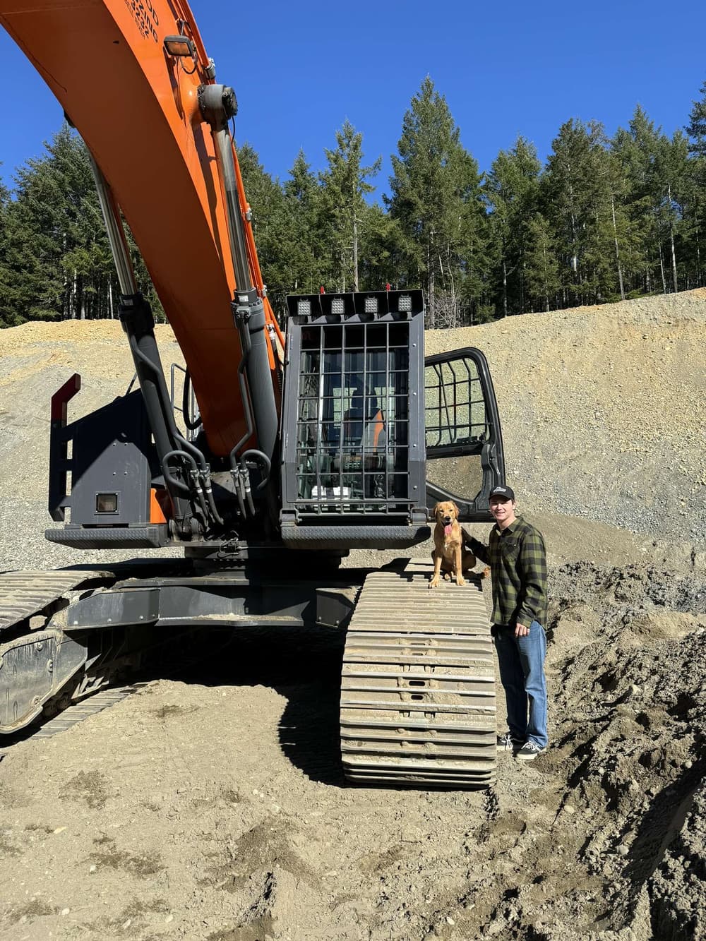 Excavator on a construction site with a man and a dog posing beside it under a clear blue sky.