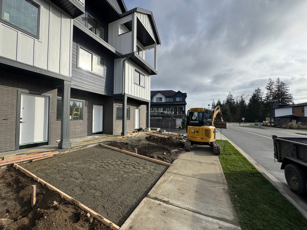 Construction site with a mini excavator and freshly laid concrete near modern buildings.