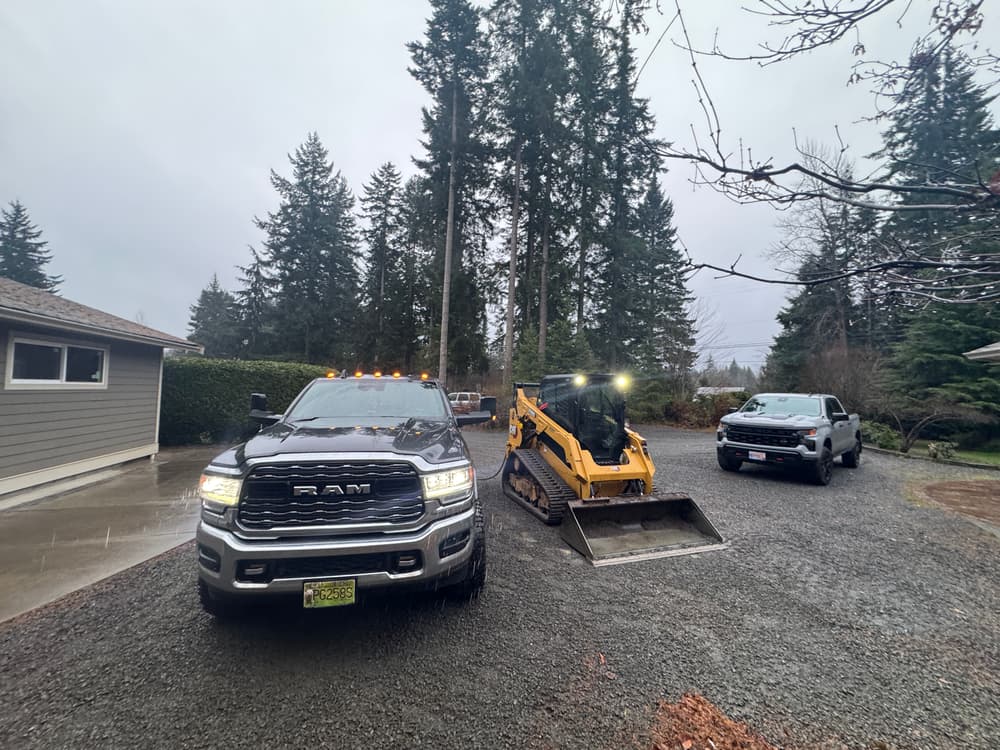 Two trucks and a compact track loader on a gravel driveway in a forested setting.